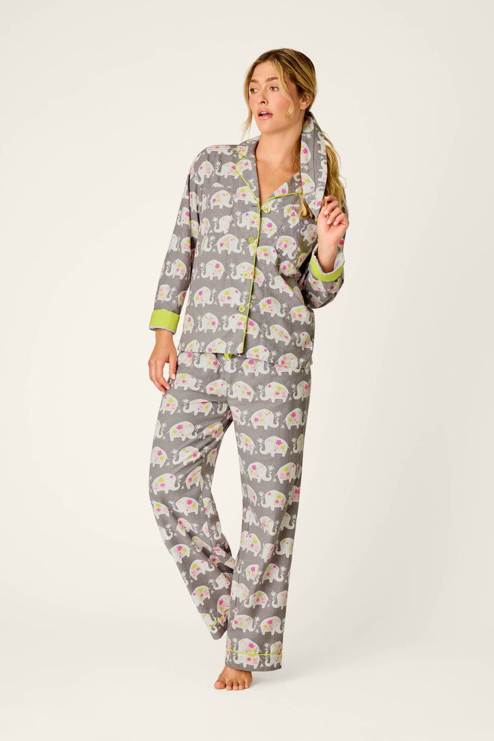 A woman models the P.J. Salvage Blooms on Parade Elephant Print 3-Piece Flannel PJ Set, featuring gray pajamas adorned with pink and white elephant designs and accented by green trim; she stands barefoot against a simple backdrop, gently touching her hair while gazing to the side.
