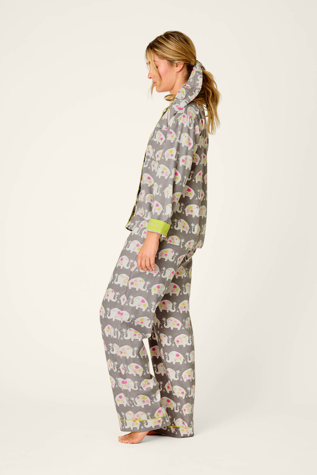 A woman stands barefoot against a plain light background, facing left in a relaxed pose, wearing the P.J. Salvage “Blooms on Parade Elephant Print” 3-Piece Flannel PJ Set decorated with colorful elephant patterns.