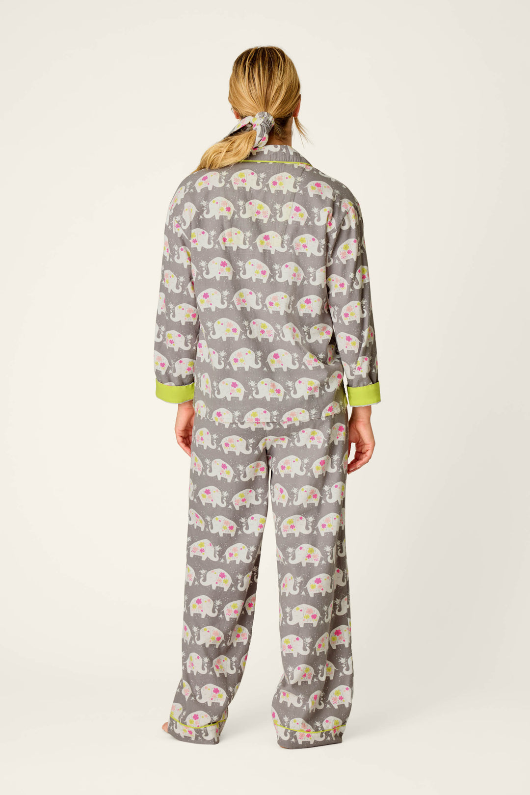 A person stands barefoot against a plain, light background, wearing the P.J. Salvage Blooms on Parade Elephant Print 3-Piece Flannel PJ Set—gray pajamas featuring a colorful elephant pattern and green cuffs. Their blonde hair is tied back with a scrunchie.