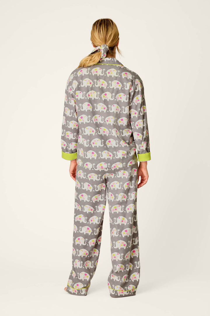 A person stands barefoot against a plain, light background, wearing the P.J. Salvage Blooms on Parade Elephant Print 3-Piece Flannel PJ Set—gray pajamas featuring a colorful elephant pattern and green cuffs. Their blonde hair is tied back with a scrunchie.