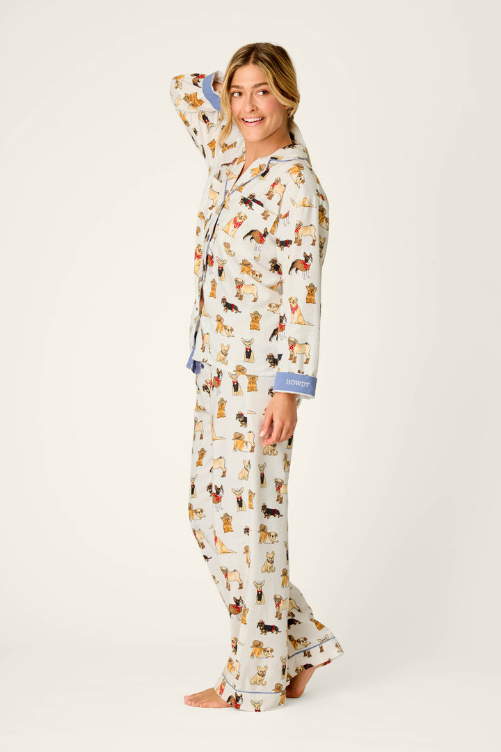 A smiling person stands barefoot, wearing the Howdy Pup-ner Dog Print 3-Piece Flannel PJ Set by P.J. Salvage—light-colored pajamas featuring playful dog illustrations and blue cuffs. With one hand behind their head, they pose against a plain background.