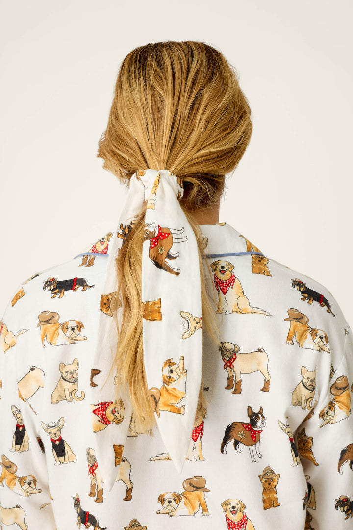 A person with blonde hair tied back with the white scarf from the P.J. Salvage Howdy Pup-ner Dog Print 3-Piece Flannel PJ Set, seen from behind. Both the scarf and their shirt display playful cartoon dog illustrations wearing hats and bandanas on a white background.