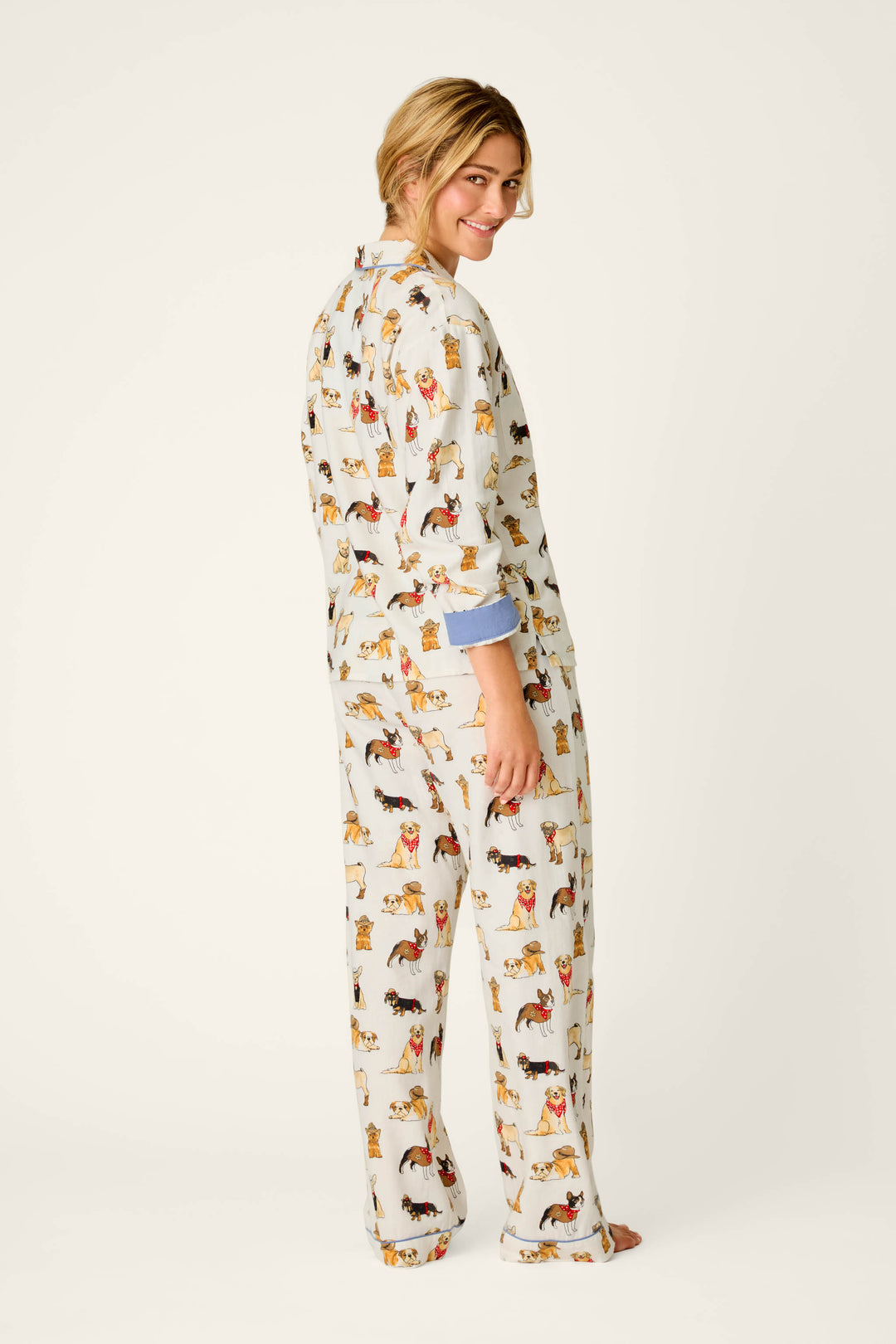 A person stands barefoot, facing backward but looking over their shoulder, wearing the P.J. Salvage Howdy Pup-ner Dog Print 3-Piece Flannel PJ Set, featuring colorful dog prints. The background is plain off-white.