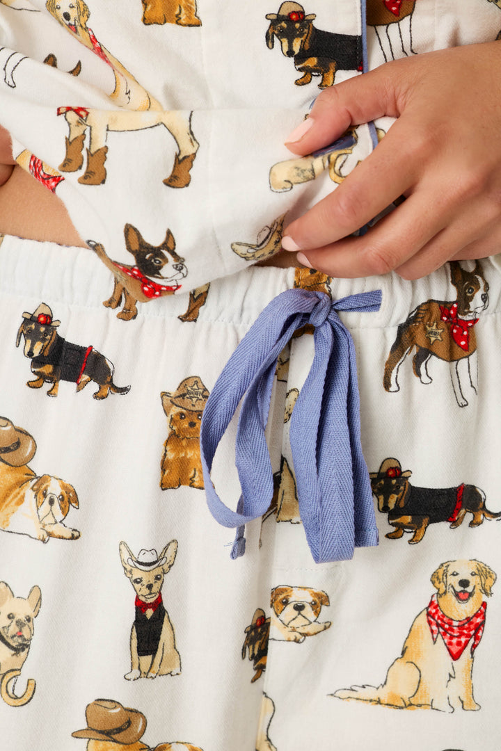 Close-up of the Howdy Pup-ner Dog Print 3-Piece Flannel PJ Set by P.J. Salvage, featuring pajama pants with a blue drawstring and a playful pattern of cartoon dogs sporting hats, bandanas, and fun accessories. A hand is shown adjusting the waistband.