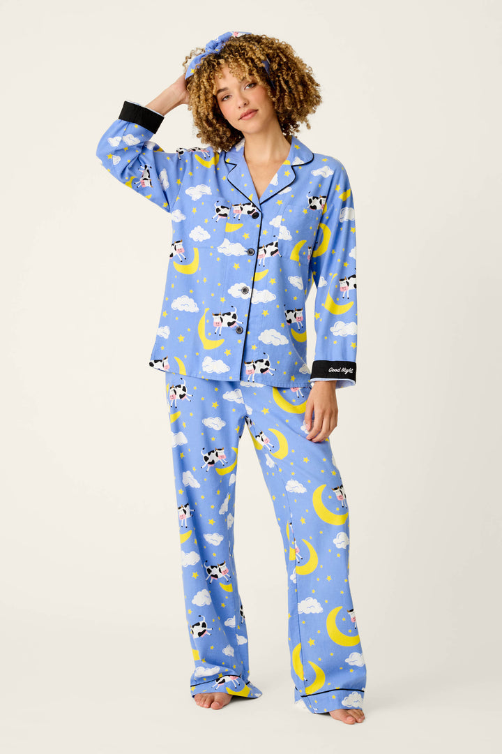 A person models the P.J. Salvage Cows Jumping Over the Moon 3-Piece Flannel PJ Set, featuring blue pajamas patterned with cows, clouds, and moons, finished with black trim, buttons, collar, and cuffs. They wear a matching headband and are barefoot while standing against a plain background with one hand resting on their head.