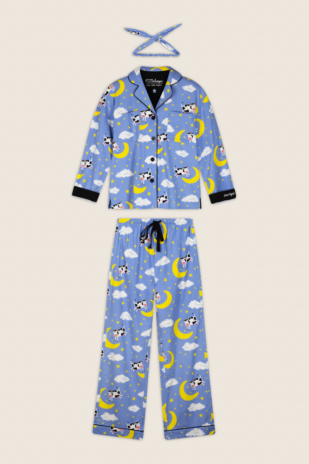 The P.J. Salvage “Cows Jumping Over the Moon” 3-Piece Flannel PJ Set features a beige background with playful cartoon cows sitting on yellow crescent moons and white clouds, accented with black trim. The set includes a long-sleeve top, pants, and a matching headband.