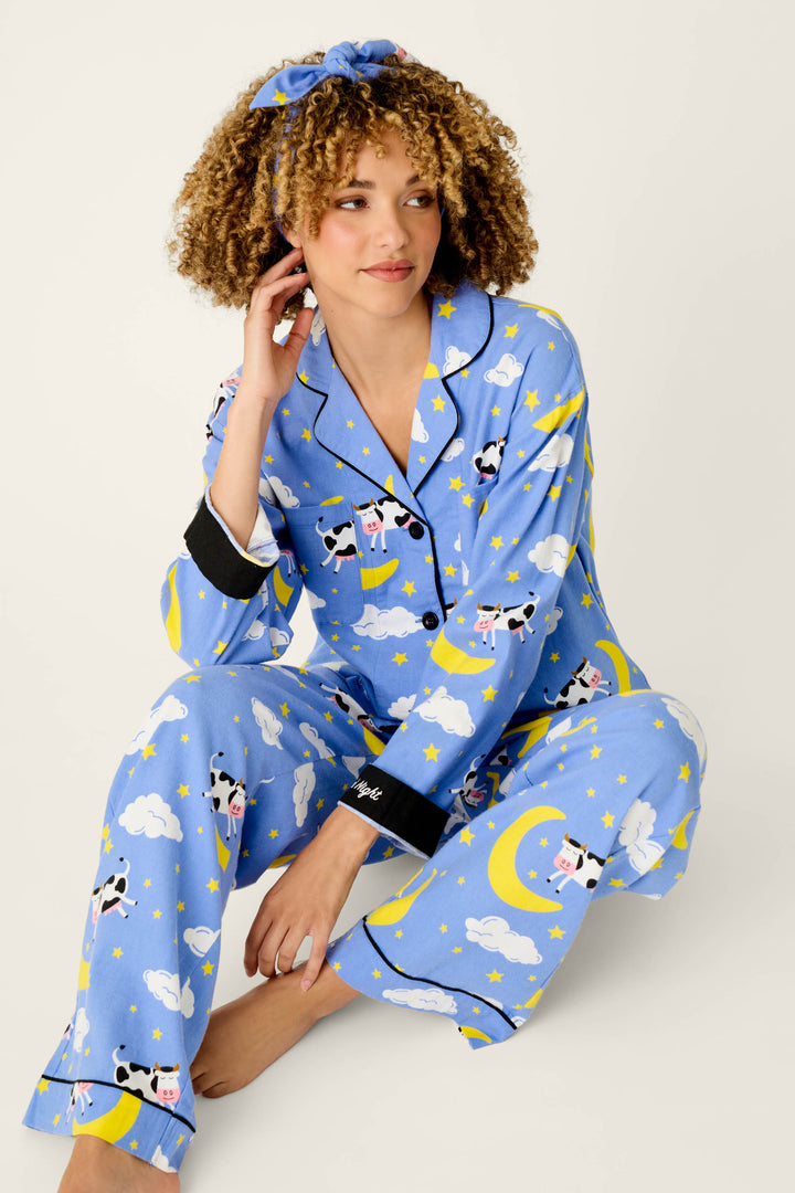 A woman with curly hair wears the P.J. Salvage Cows Jumping Over the Moon 3-Piece Flannel PJ Set, featuring coordinating blue pajamas and a matching headband adorned with whimsical cow, cloud, and moon motifs. Sitting on the floor with one hand in her hair, she gazes off to the side with a gentle smile.