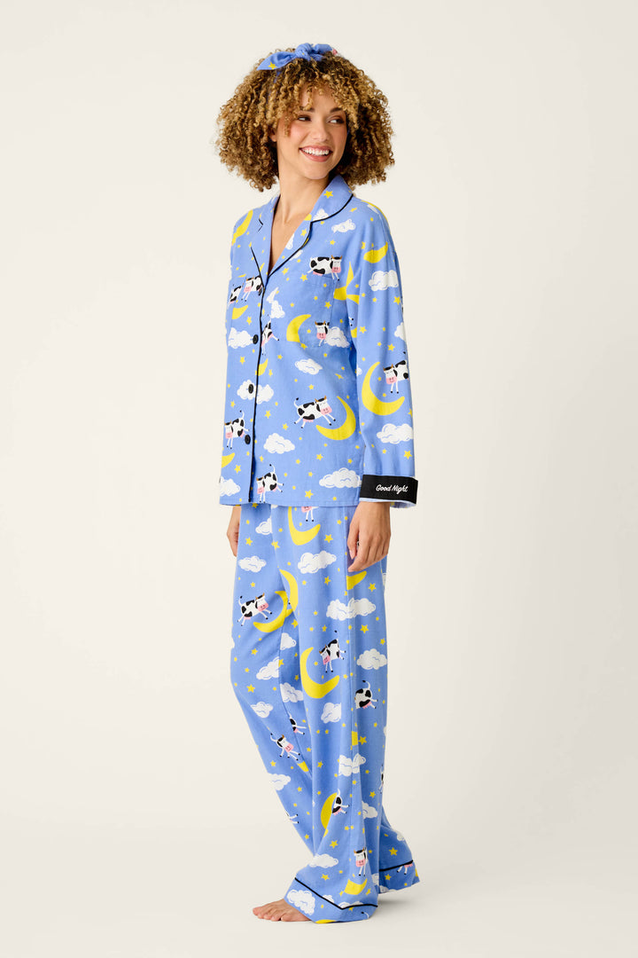 A smiling person wears the P.J. Salvage Cows Jumping Over the Moon 3-Piece Flannel PJ Set, featuring a playful cow and moon pattern on blue fabric, with a matching scrunchie and black cuffs embroidered with “Good night,” standing barefoot against a plain background.