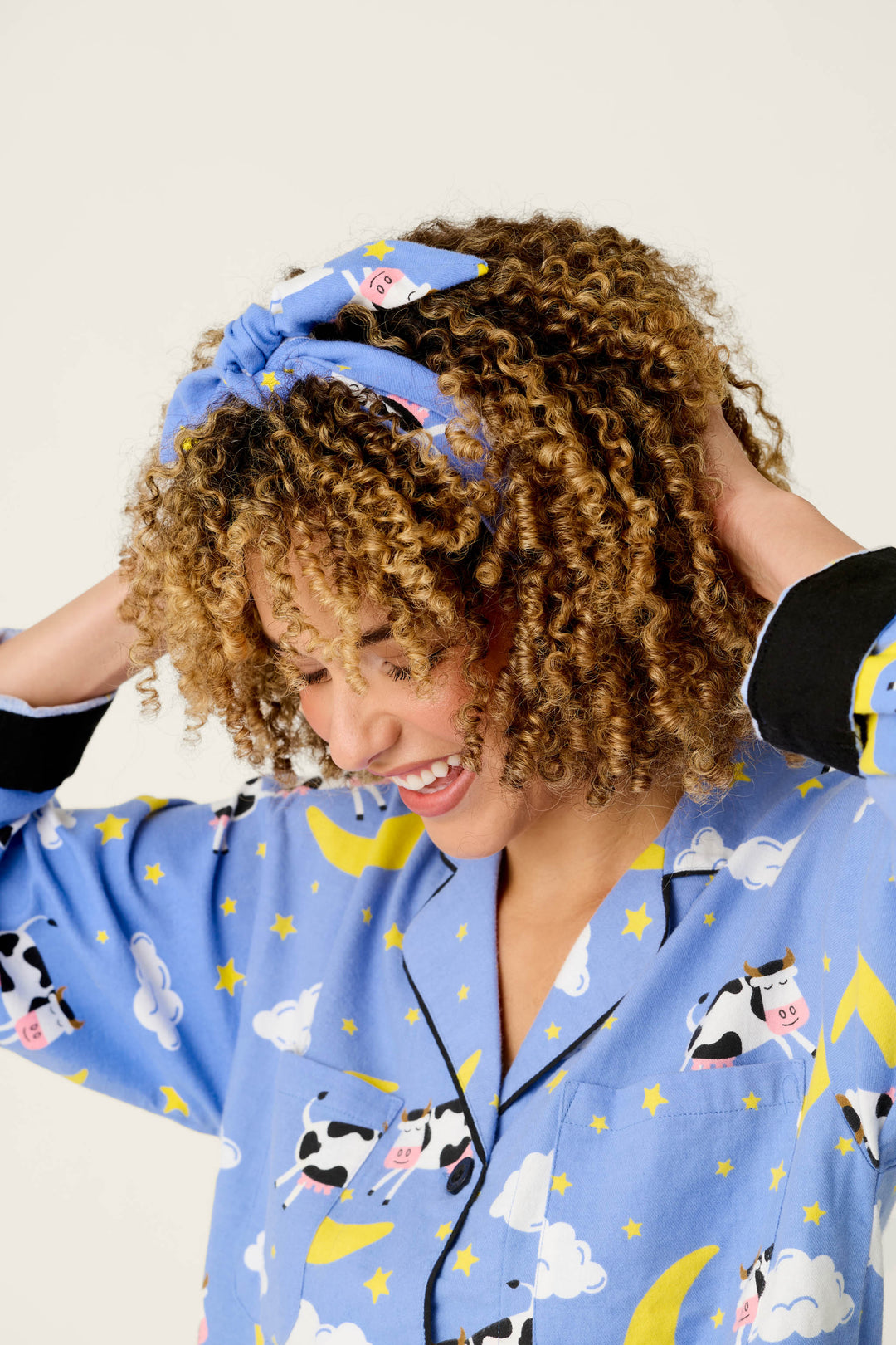 A smiling person with curly hair wears the P.J. Salvage Cows Jumping Over the Moon 3-Piece Flannel PJ Set, featuring blue pajamas with cow, moon, and star prints and a matching headband. They look down while touching their hair with both hands.