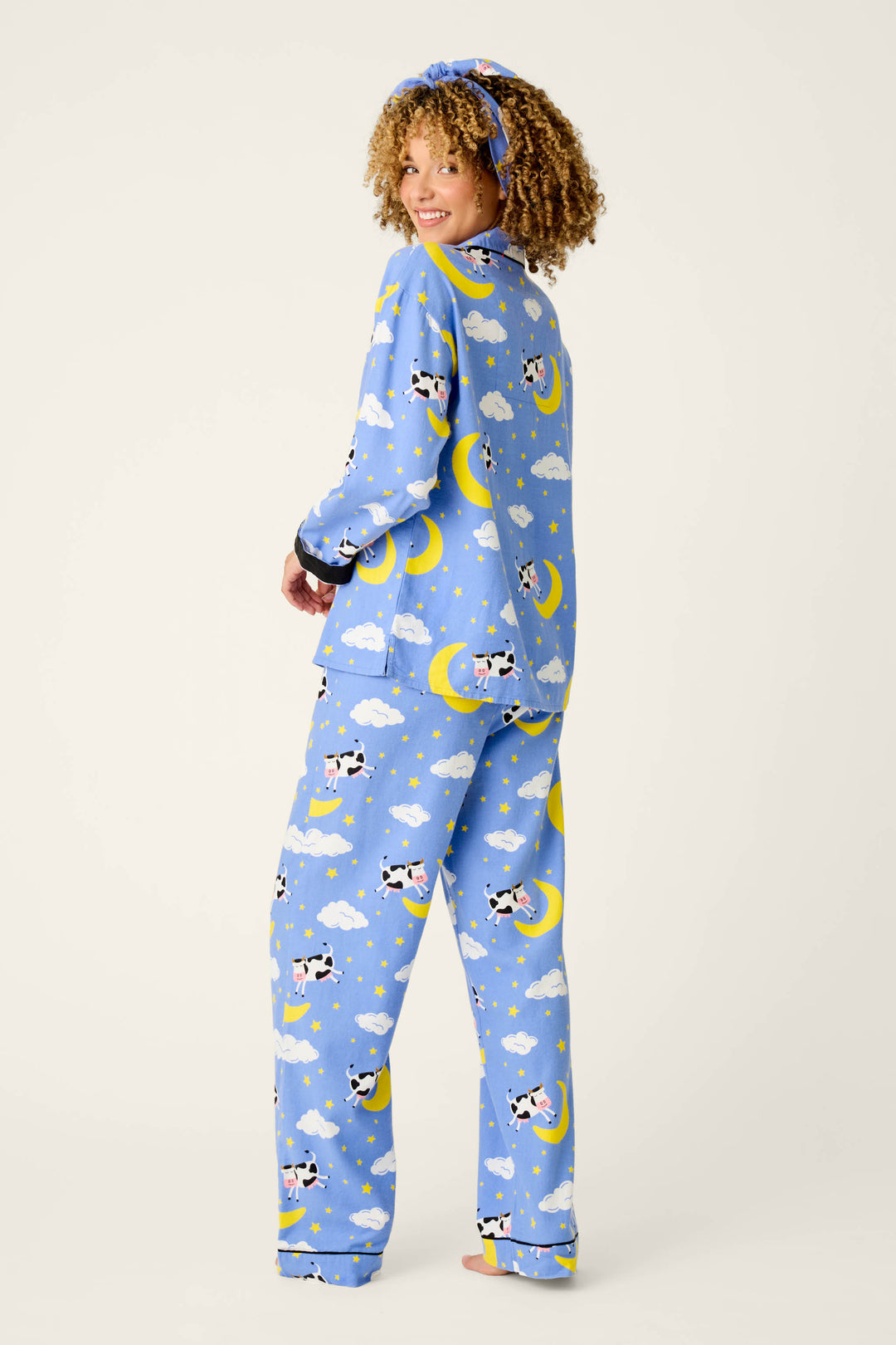 A woman smiles over her shoulder while wearing the P.J. Salvage Cows Jumping Over the Moon 3-Piece Flannel PJ Set—featuring whimsical cow, moon, and cloud patterns—and a matching headband. She stands barefoot against a plain background.