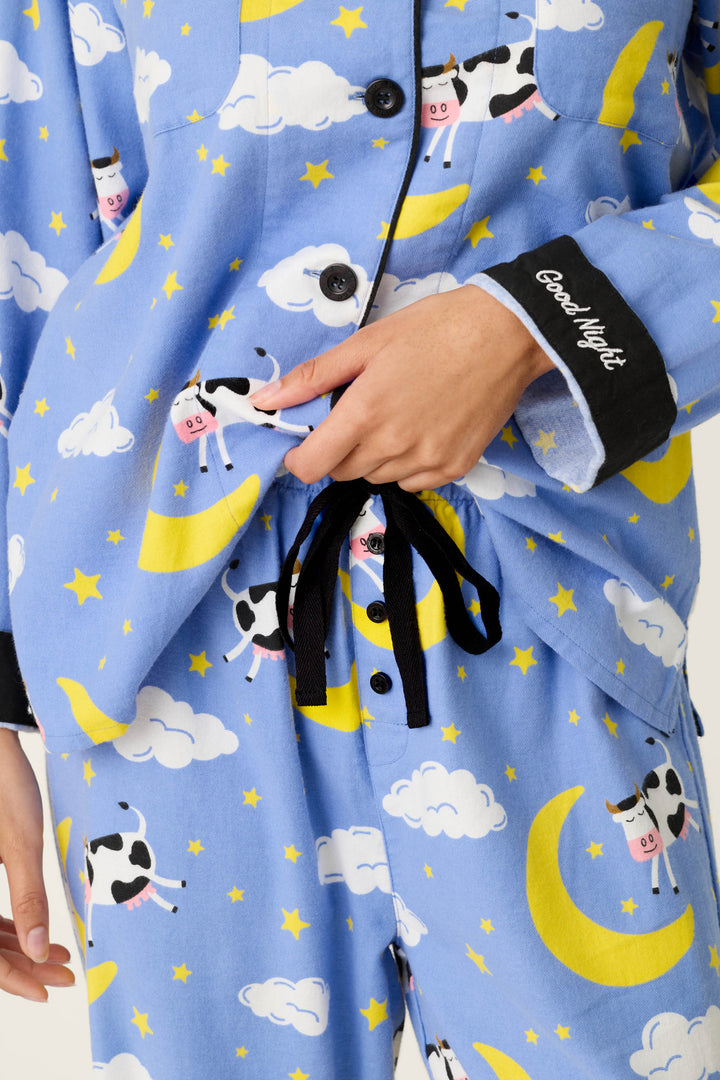 A person is shown adjusting the black drawstring waist of the pants from the P.J. Salvage Cows Jumping Over the Moon 3-Piece Flannel PJ Set, which features blue fabric with yellow moons, white clouds, stars, and playful cartoon cows. The pajama sleeve is printed with “Good Night” in white script.