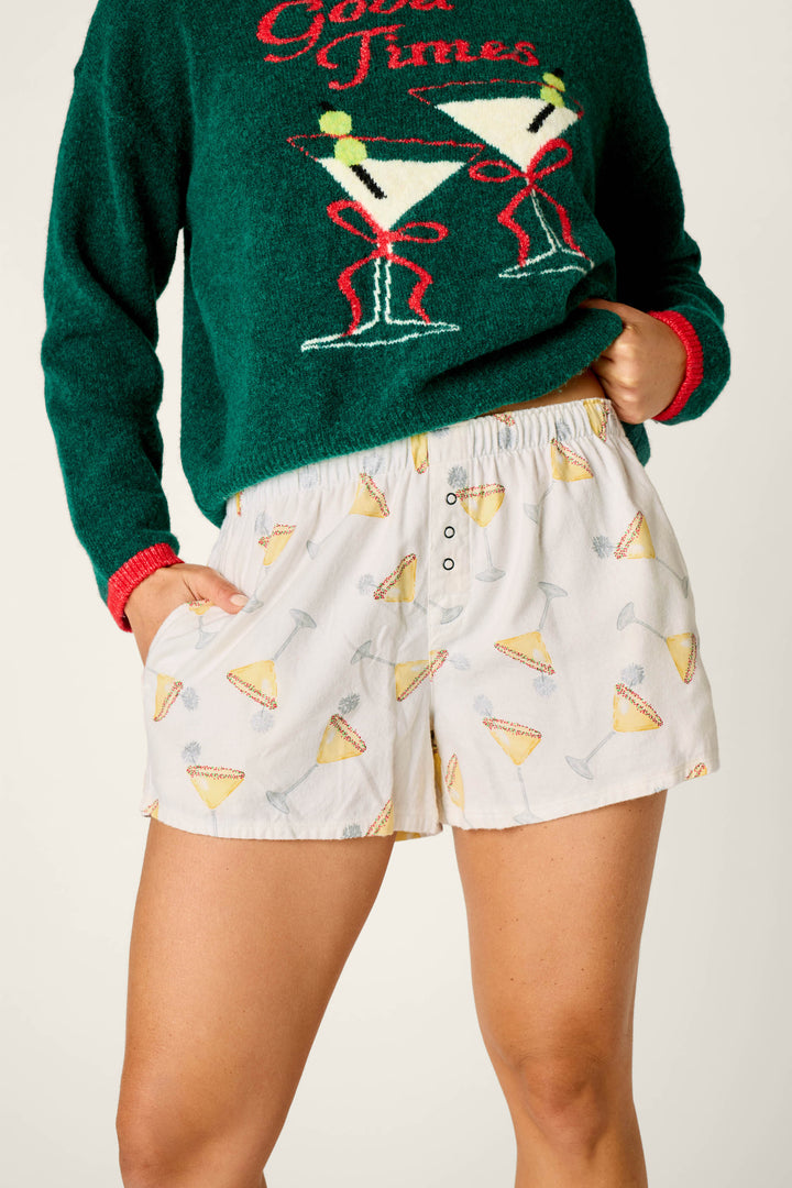 A person wearing the P.J. Salvage Happy Hour Cocktail Print Flannel Pajama Shorts with a green sweater featuring two martini glasses and red text, standing with hands in their pockets.