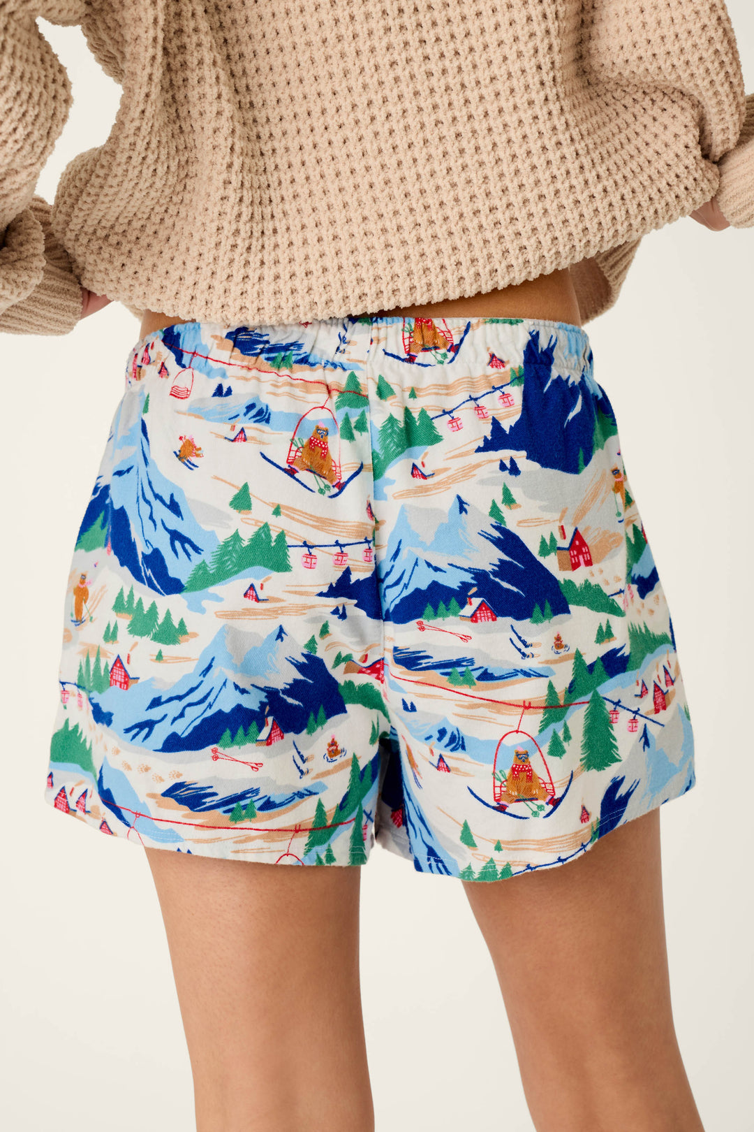 A person wearing P.J. Salvage Ski Ya Later Skiing Bears Print Flannel Pajama Shorts and a beige waffle-knit sweater. The image is cropped to show only the shorts and part of the sweater.
