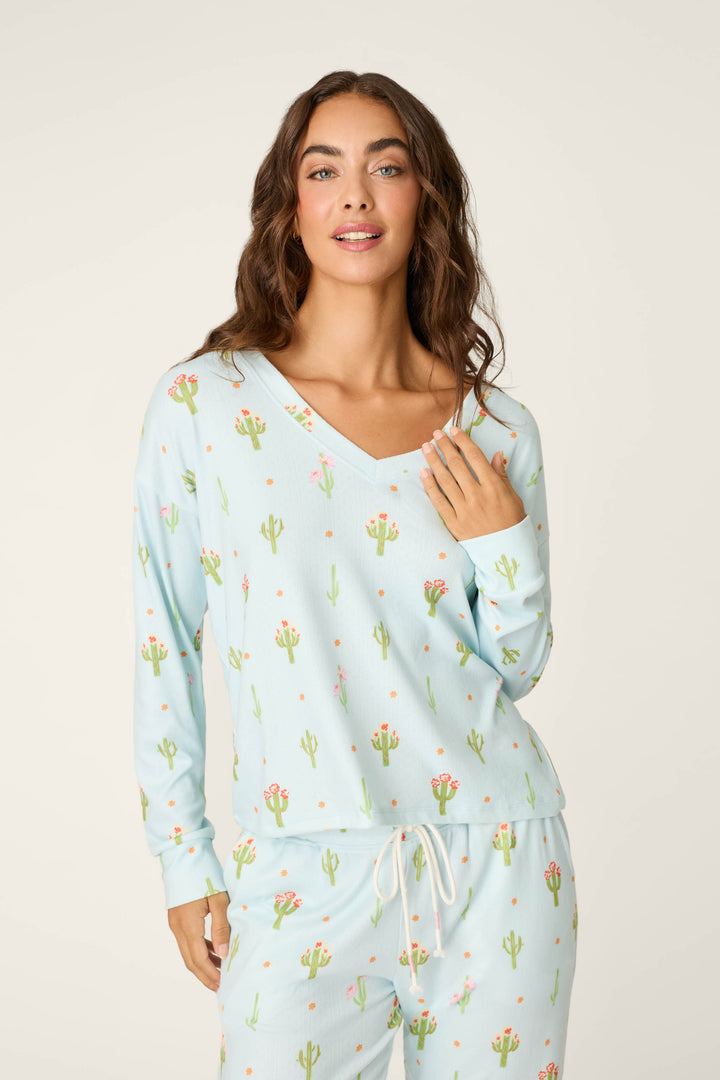 A woman with wavy brown hair wears the P.J. Salvage Feelin' Prickly Cactus Blooms Long Sleeve Top, a light blue pajama set with cactus print, perfect for cozy nights, standing against a plain light background.