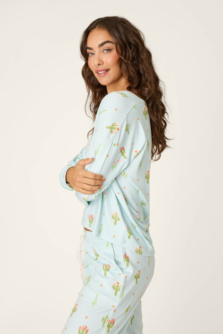 A woman with long wavy brown hair models the P.J. Salvage Feelin' Prickly Cactus Blooms Long Sleeve Top, featuring a light blue cactus print, perfect for cozy nights. She stands sideways, arms crossed, smiling gently against a plain background.