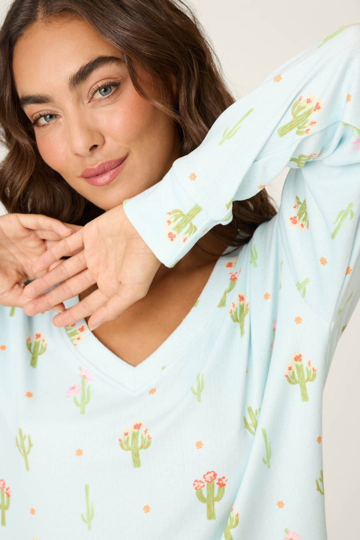 A woman with wavy brown hair smiles softly, raising her arms near her face, wearing the Feelin' Prickly Cactus Blooms Long Sleeve Top by P.J. Salvage—ideal for cozy nights in.
