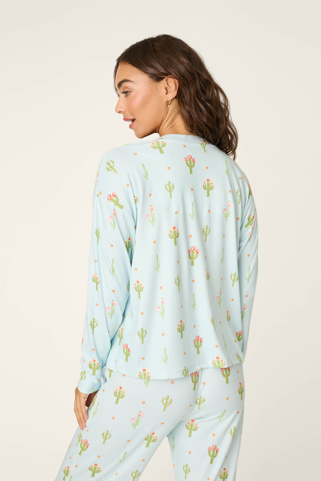 A woman with wavy brown hair wears the P.J. Salvage Feelin' Prickly Cactus Blooms Long Sleeve Top and light blue pajamas, looking back over her shoulder against a cream background—an ideal choice for cozy nights.