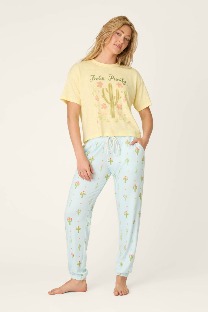A woman stands barefoot in a yellow Feelin Prickly T-shirt with cacti and flower graphics, and P.J. Salvage Feelin' Prickly Cactus Blooms Banded Pants decorated with small cactus designs, posing against an off-white background.