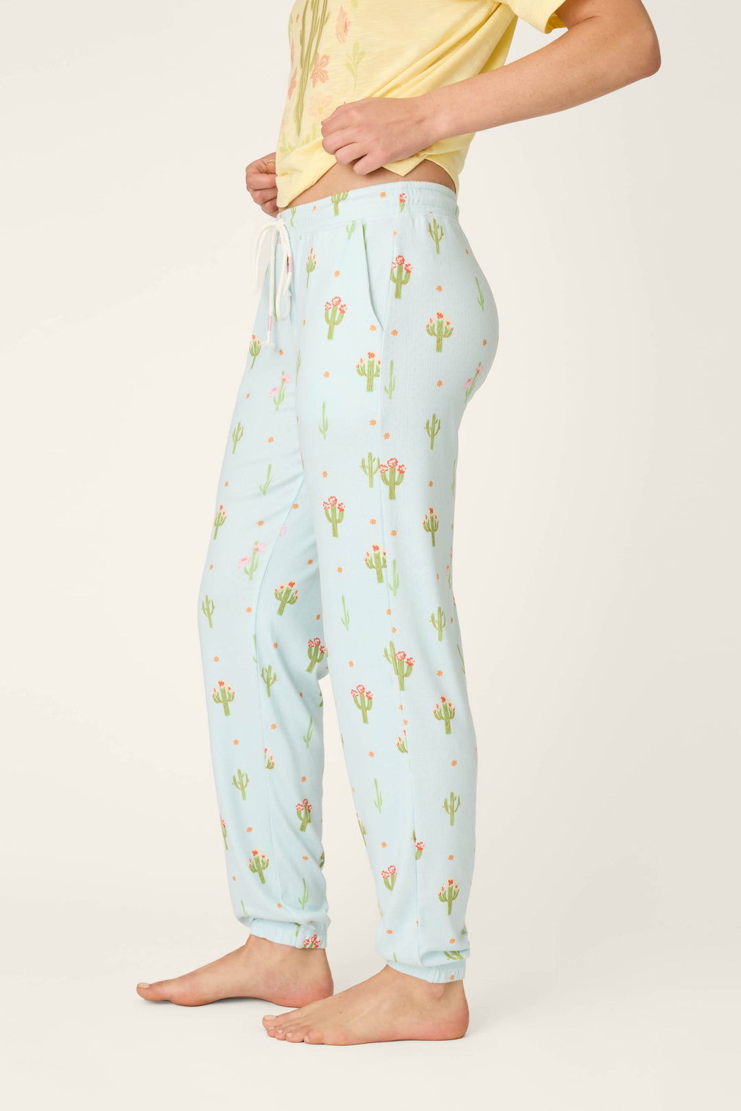 A person stands barefoot in P.J. Salvage Feelin' Prickly Cactus Blooms Banded Pants—relaxed fit, light blue with a cactus print—paired with a yellow T-shirt, shown from the side against a plain background. Perfect for cozy nights.