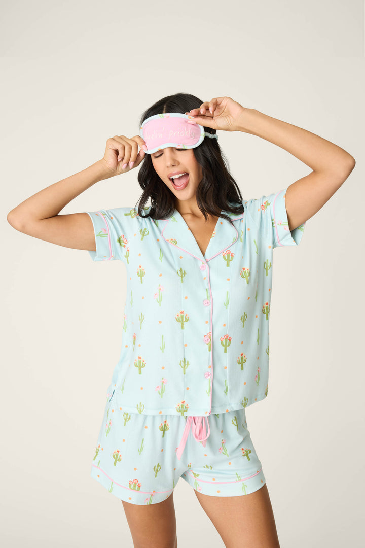 A woman poses cheerfully in the P.J. Salvage Feelin' Prickly Cactus Blooms 3-Piece Short PJ Set, wearing a pink sleep mask and holding it up with both hands, one eye closed and mouth open.