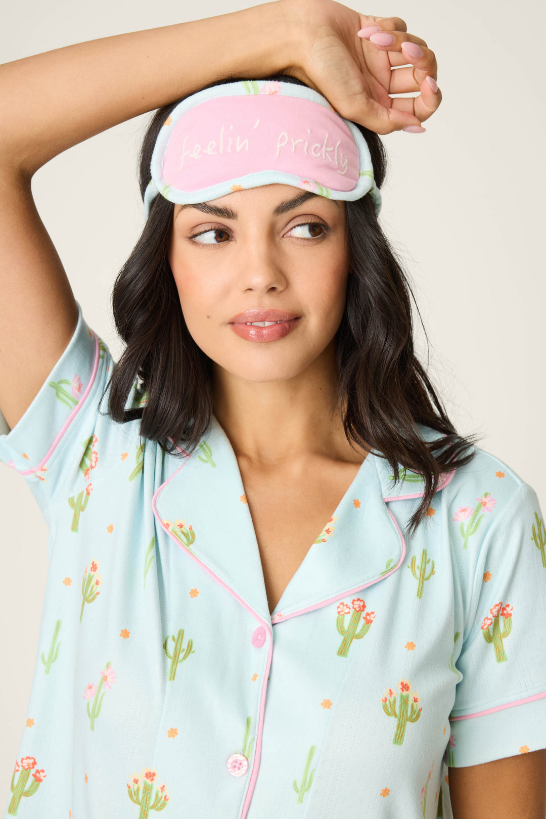 A woman in the Feelin' Prickly Cactus Blooms 3-Piece Short PJ Set by P.J. Salvage, featuring pastel blue cactus print shorts, adjusts a pink and blue sleep mask with "feelin prickly" text. She has wavy dark hair and looks to the side.