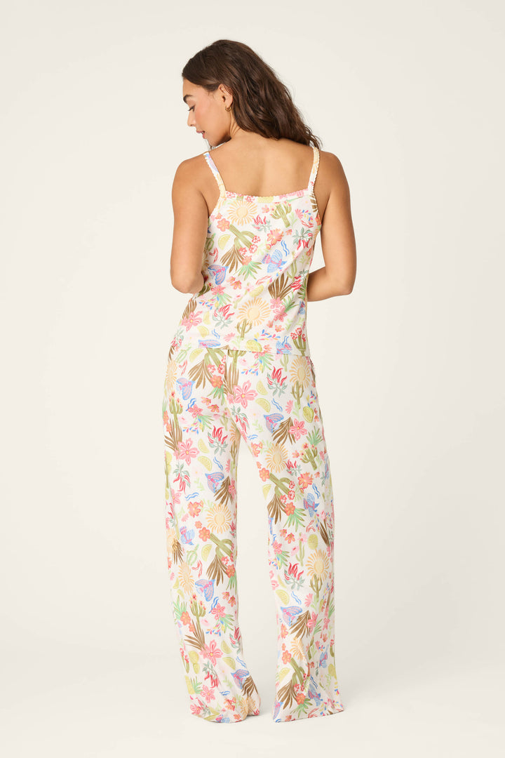 A woman faces away, wearing the Feelin' Prickly Desert Dreams Long PJ Set by P.J. Salvage—featuring a sleeveless top with thin straps and loose pants in a pink, green, blue, and beige desert print against a light background.