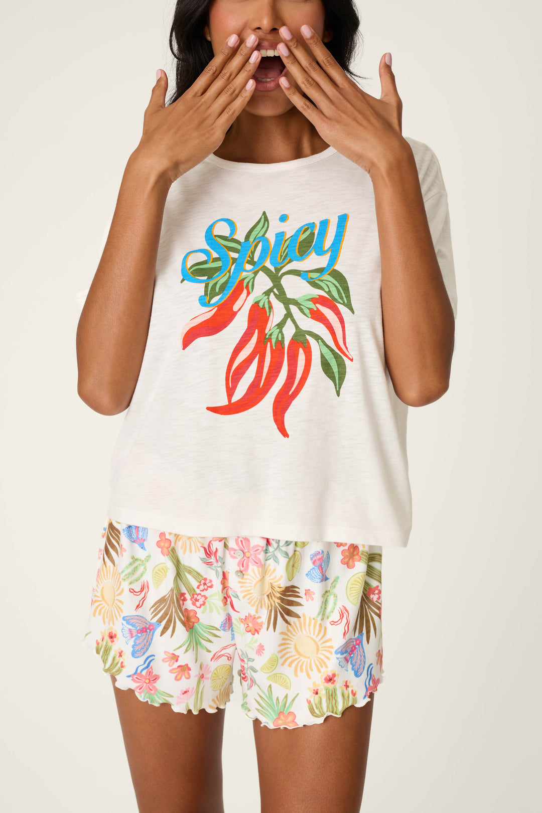 A woman playfully poses against a light background, wearing the P.J. Salvage Feelin’ Prickly Spicy Chili Graphic T-Shirt and floral shorts. She holds her hands by her mouth, showcasing the shirt's bold chili graphic.