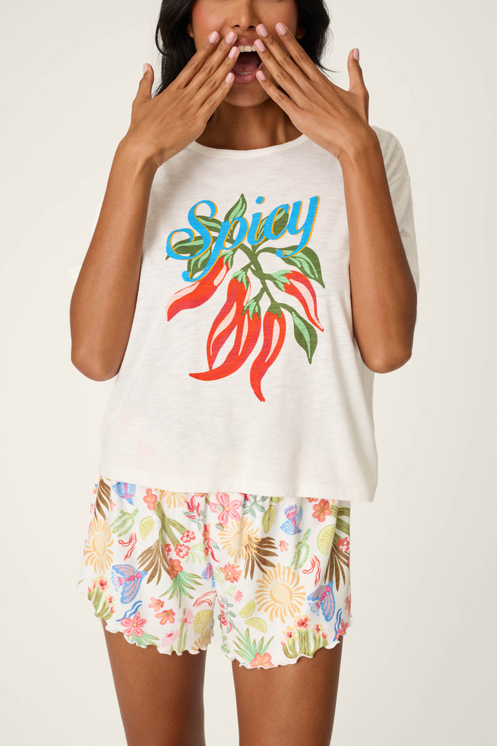 A woman playfully poses against a light background, wearing the P.J. Salvage Feelin’ Prickly Spicy Chili Graphic T-Shirt and floral shorts. She holds her hands by her mouth, showcasing the shirt's bold chili graphic.