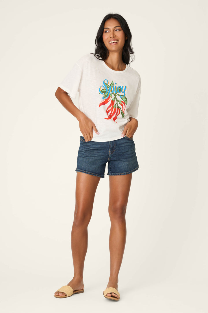 A woman smiles in a Feelin’ Prickly Spicy Chili Graphic T-Shirt by P.J. Salvage, paired with dark denim shorts and tan slide sandals, posed against a plain light background.