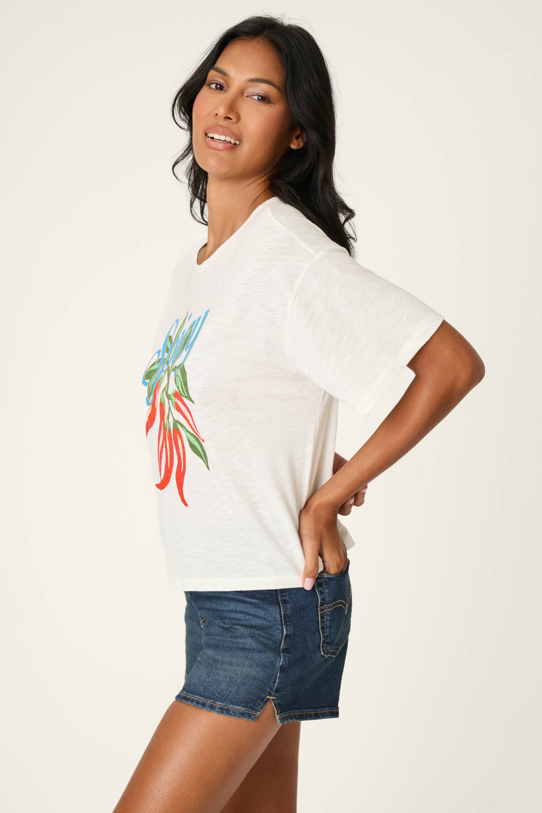 A woman with long dark hair models the P.J. Salvage Feelin’ Prickly Spicy Chili Graphic T-Shirt, paired with denim shorts, posing sideways and smiling at the camera against a plain background.
