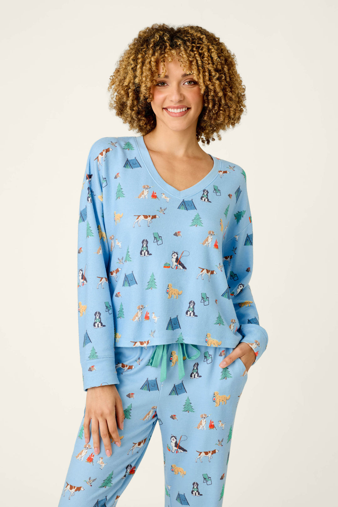 A smiling person with curly hair is wearing the P.J. Salvage “Happy Campawr Dogs Camping Print” long sleeve top, featuring light blue fabric with playful camping-themed designs—tents, trees, and dogs—standing against a plain background.
