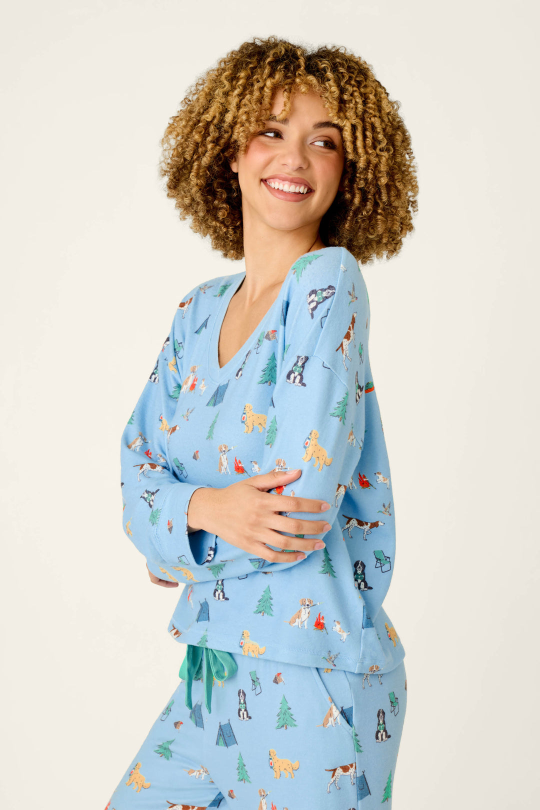 A woman with curly hair smiles while wearing the P.J. Salvage Happy Campawr Dogs Camping Print Long Sleeve Top—light blue pajamas adorned with playful dog and tree motifs—standing against a plain background.