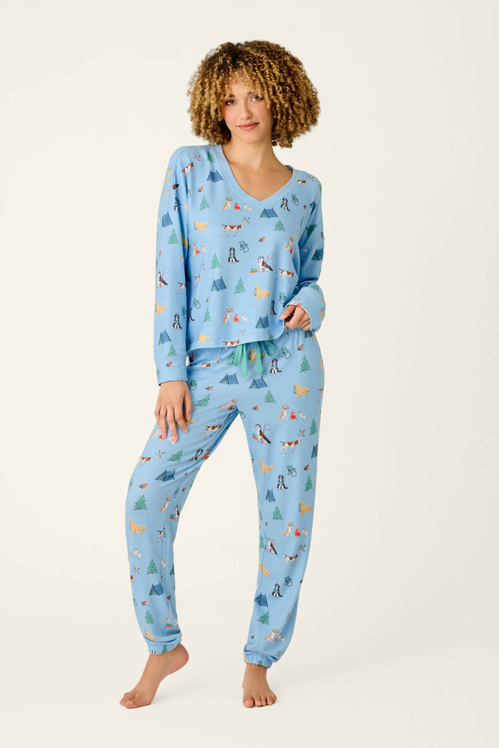 A person with curly hair wears the P.J. Salvage Happy Campawr Dogs Camping Print Long Sleeve Top in light blue, featuring a fun, colorful animal and tree pattern, standing barefoot against a plain white background.
