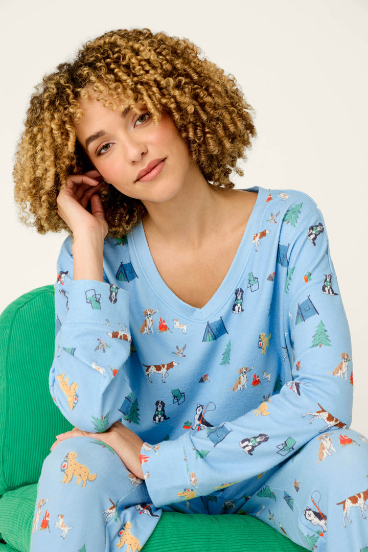 A woman with curly hair sits on a green chair, wearing the P.J. Salvage Happy Campawr Dogs Camping Print Long Sleeve Top. She rests her head on her hand and looks at the camera with a slight smile.