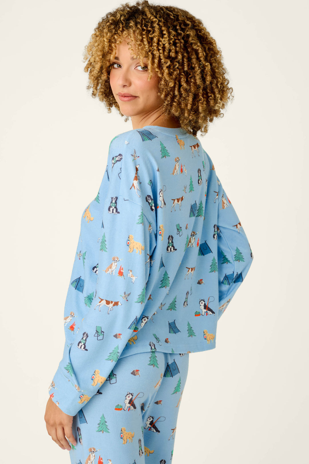 A woman with curly hair is wearing the P.J. Salvage Happy Campawr Dogs Camping Print Long Sleeve Top, featuring colorful cartoon dogs, tents, and trees on a light blue background. She stands sideways, smiling slightly as she looks back at the camera against a plain light backdrop.