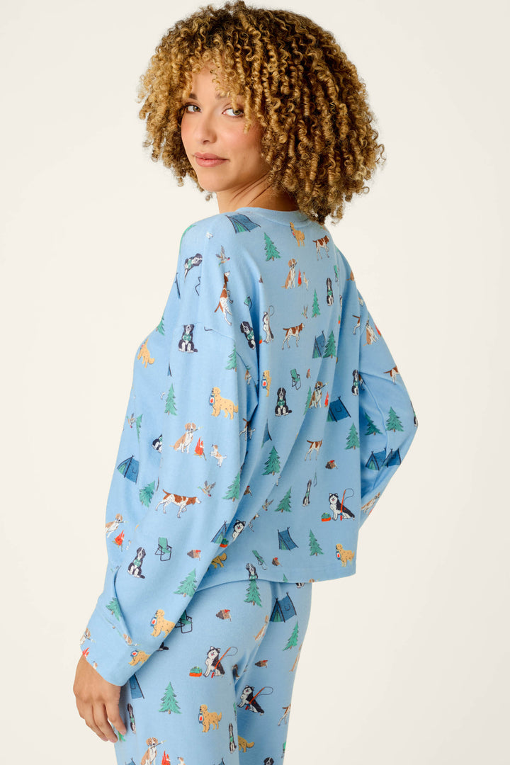 A woman with curly hair is wearing the P.J. Salvage Happy Campawr Dogs Camping Print Long Sleeve Top, featuring colorful cartoon dogs, tents, and trees on a light blue background. She stands sideways, smiling slightly as she looks back at the camera against a plain light backdrop.