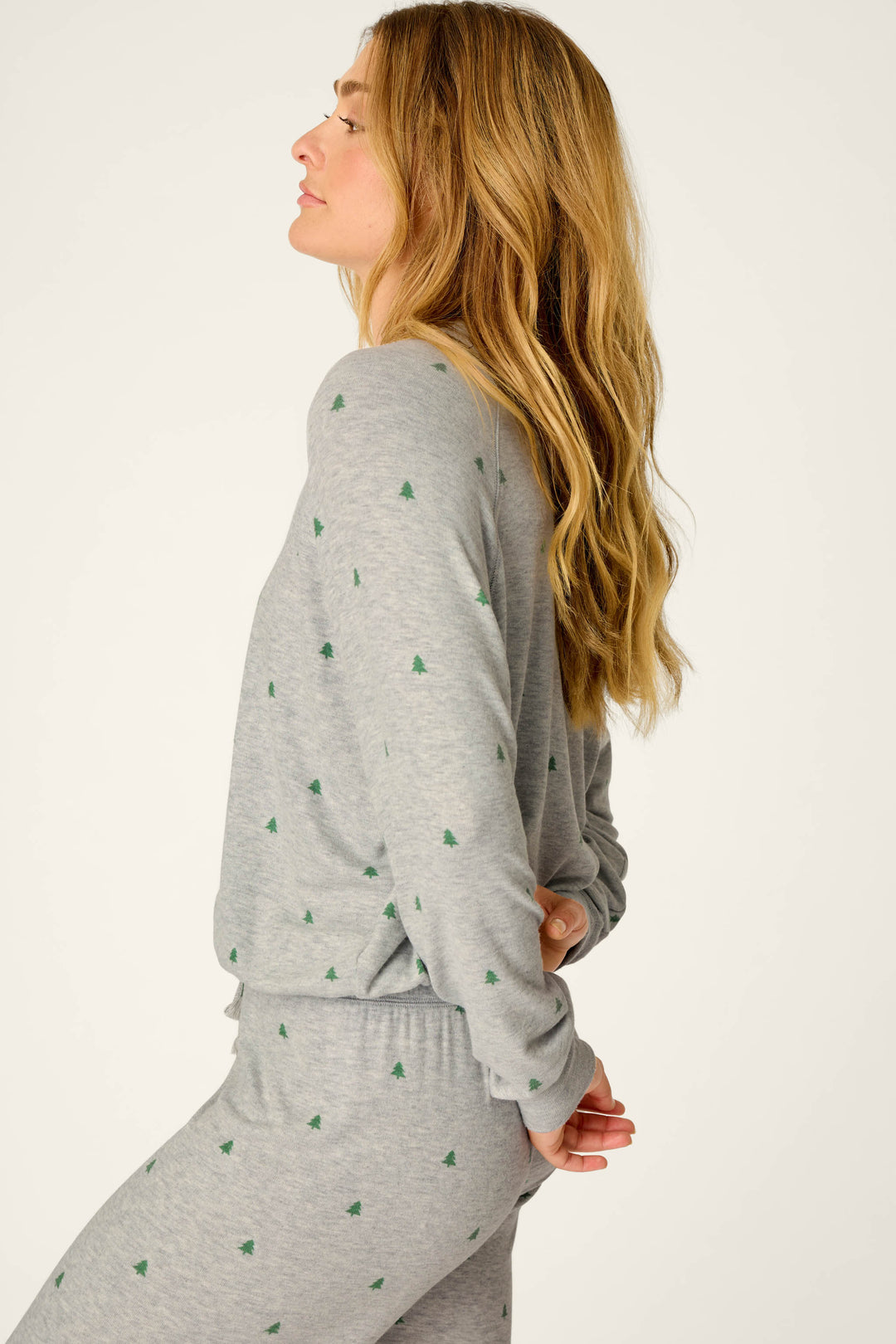 A woman with long, wavy blonde hair stands in profile against a plain light background, wearing the P.J. Salvage Happy Campawr Tree Print Long Sleeve Top and matching grey pajama bottoms adorned with small green tree prints, her hands clasped behind her back.