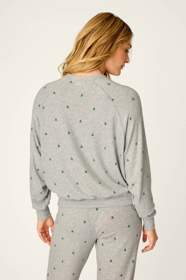 A person with long, wavy blonde hair stands with their back to the camera against an off-white background, wearing the P.J. Salvage Happy Campawr Tree Print Long Sleeve Top and matching pants featuring a small green tree pattern.