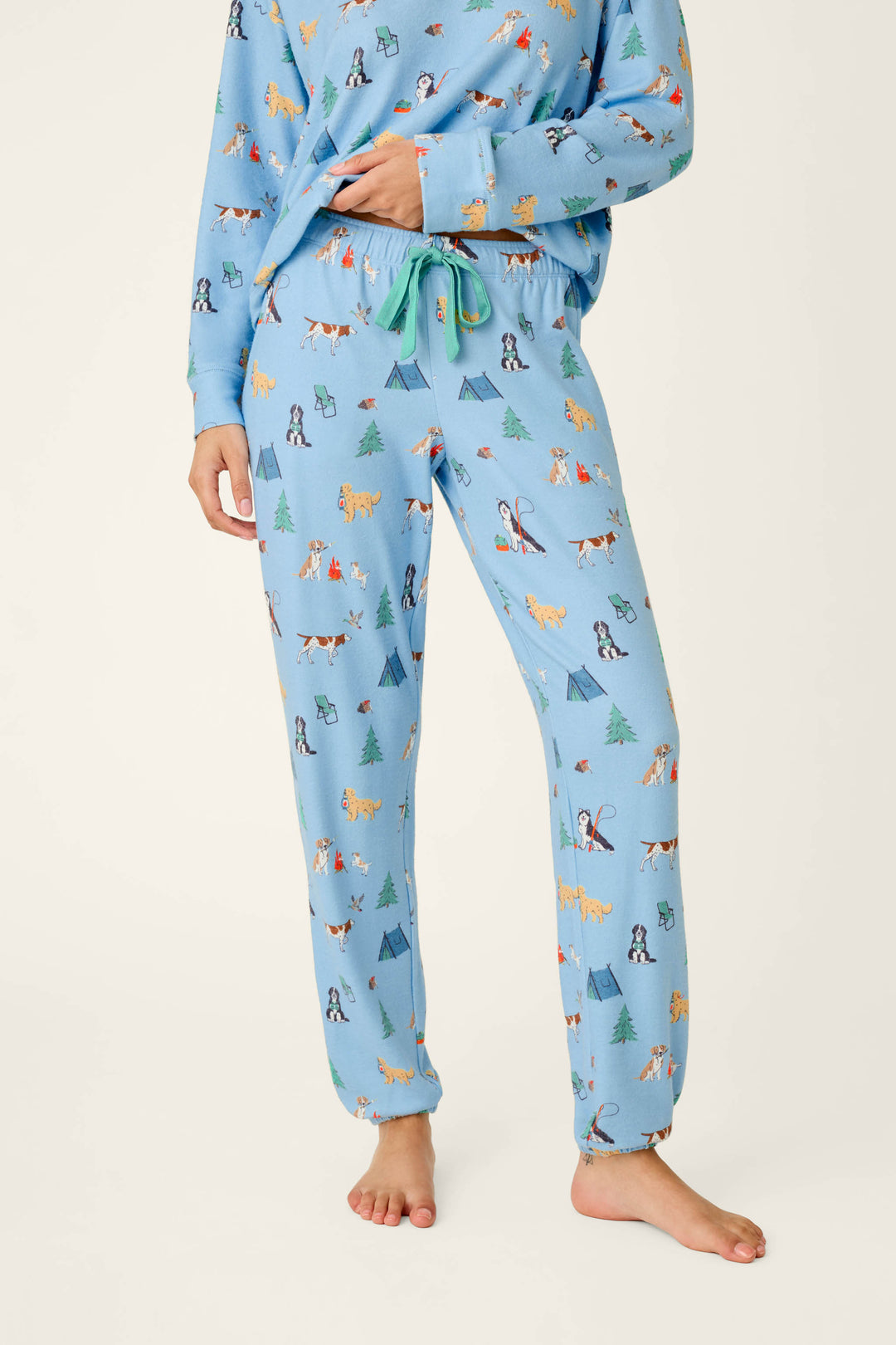 A person wears the P.J. Salvage Happy Campawr Dogs Camping Print Jogger Pajama Pants, featuring a light blue background with playful camping-themed prints of dogs, tents, trees, and campfires. The matching pajama top is partially visible as the barefoot individual stands on a light background.