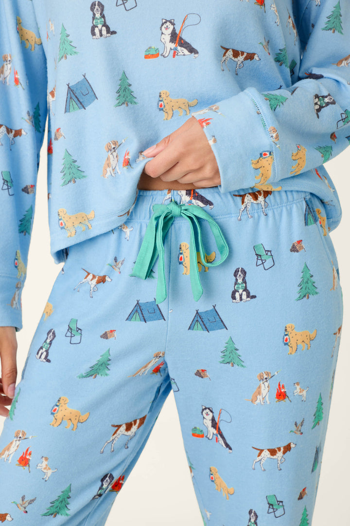 A person wearing the P.J. Salvage Happy Campawr Dogs Camping Print Jogger Pajama Pants in light blue, featuring playful dogs, tents, trees, campfires, and backpacks in a cheerful camping-themed print. The individual is shown holding the hem of their shirt.