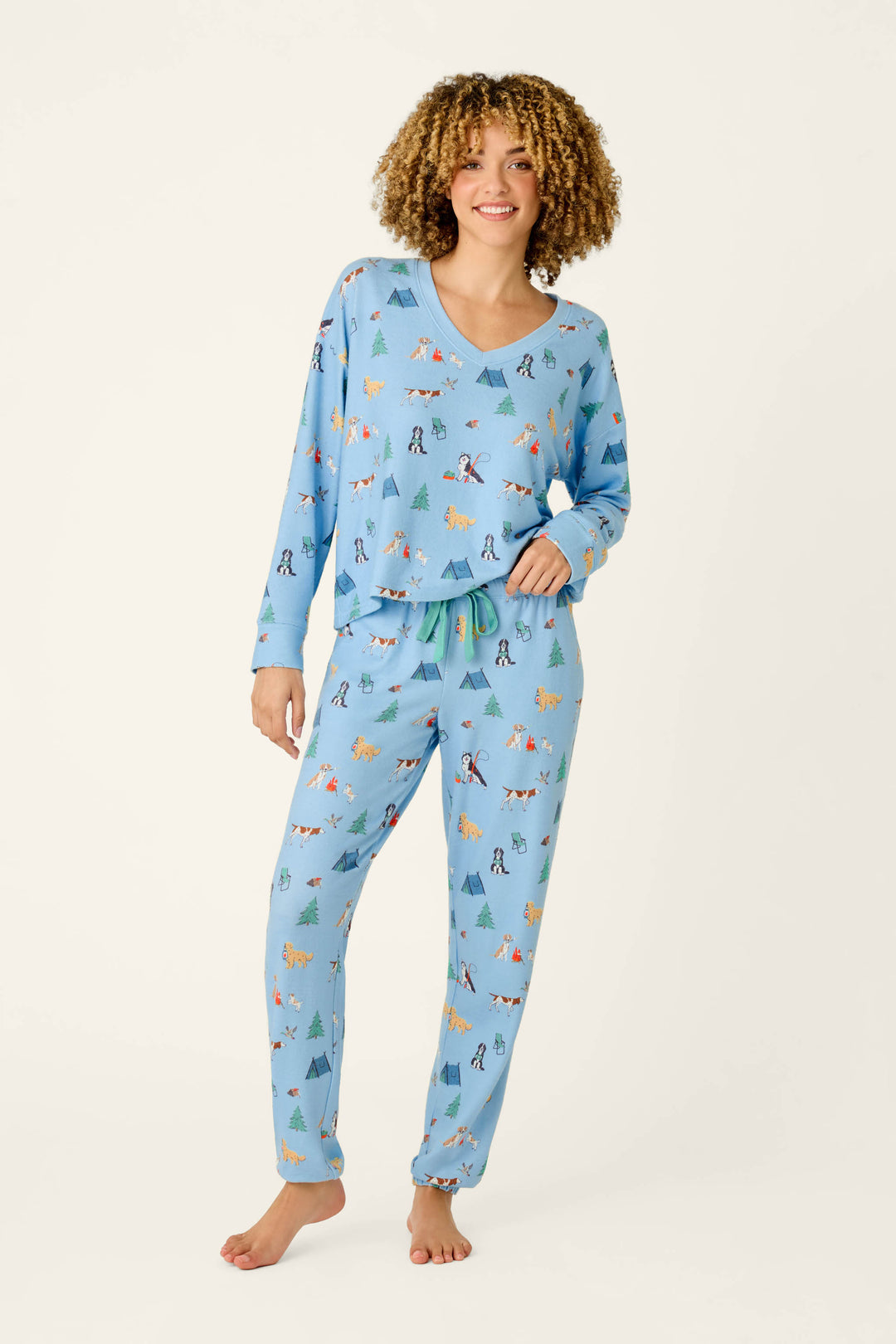 A person stands barefoot, smiling, wearing P.J. Salvage Happy Campawr Dogs Camping Print Jogger Pajama Pants.