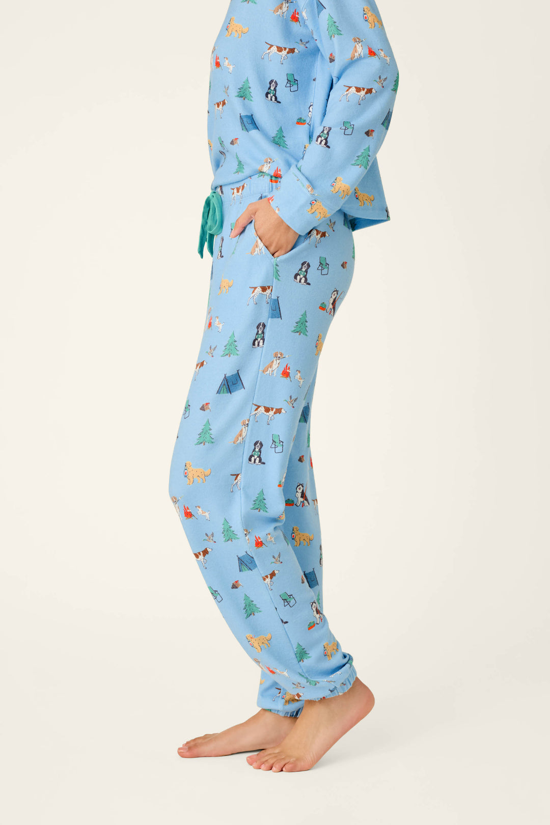 A person stands barefoot, wearing P.J. Salvage Happy Campawr Dogs Camping Print Jogger Pajama Pants in light blue, featuring a playful pattern of dogs, tents, and trees. The person’s head and upper body are not visible.