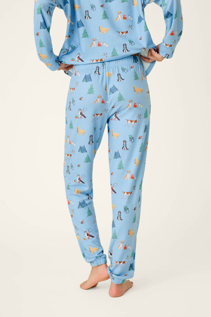A person is shown from the lower back and legs, standing barefoot on a light background, wearing P.J. Salvage Happy Campawr Dogs Camping Print Jogger Pajama Pants featuring colorful animal and tree prints; hands rest on their hips.