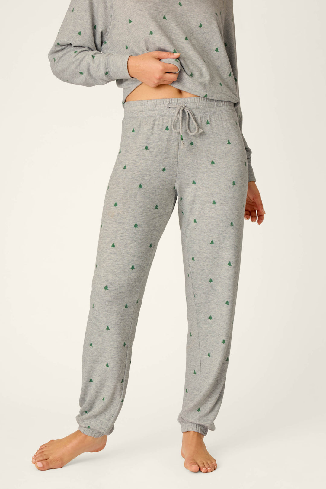 A person is standing barefoot on a light background, wearing P.J. Salvage Happy Campawr Tree Print Jogger Pants and a matching long-sleeve top featuring small green tree patterns.