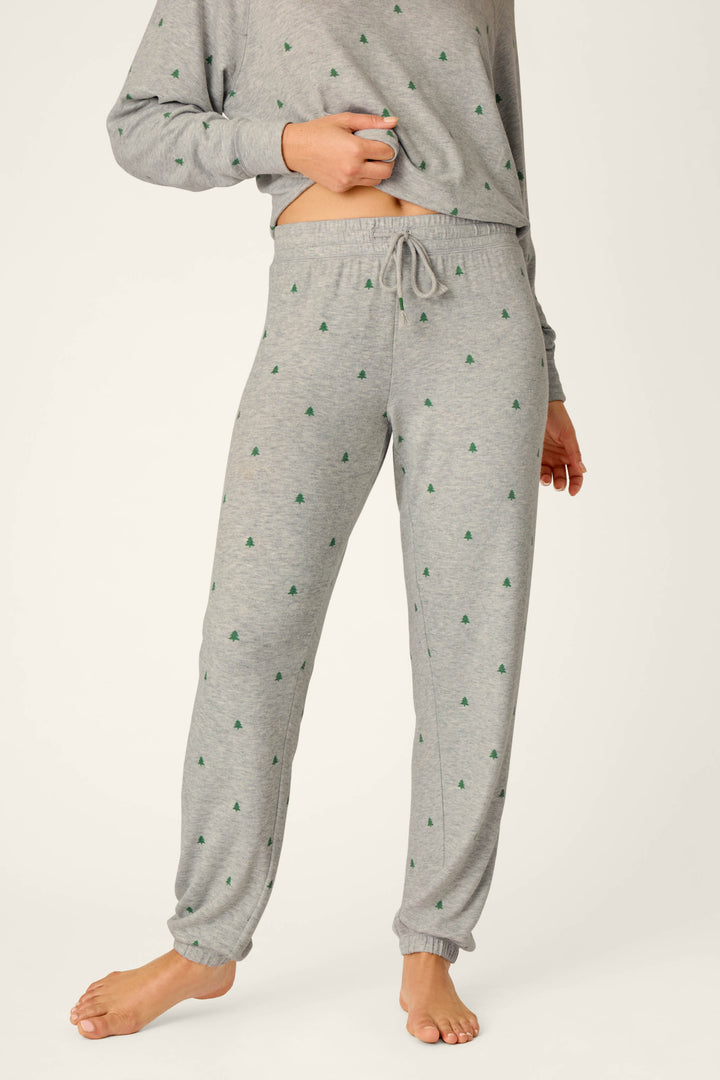 A person is standing barefoot on a light background, wearing P.J. Salvage Happy Campawr Tree Print Jogger Pants and a matching long-sleeve top featuring small green tree patterns.