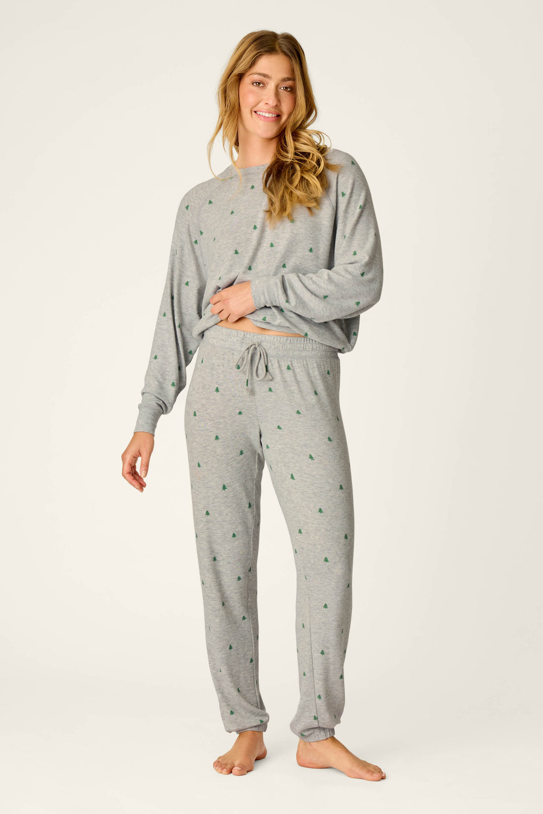 A woman stands barefoot against a plain background, smiling with one hand on her hip. She wears the P.J. Salvage Happy Campawr Tree Print Jogger Pants, featuring a drawstring waist and small green tree patterns.