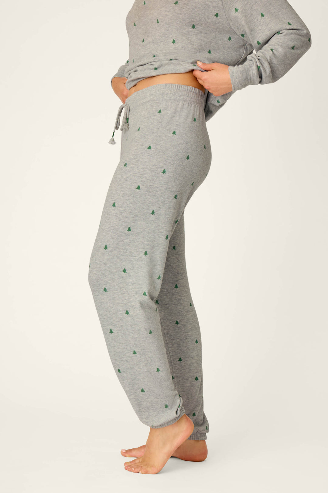 A person is shown from the waist down, barefoot, wearing P.J. Salvage’s Happy Campawr Tree Print Jogger Pants in light grey with small green triangle patterns, paired with a matching sweatshirt and lifting the top slightly for a relaxed look.