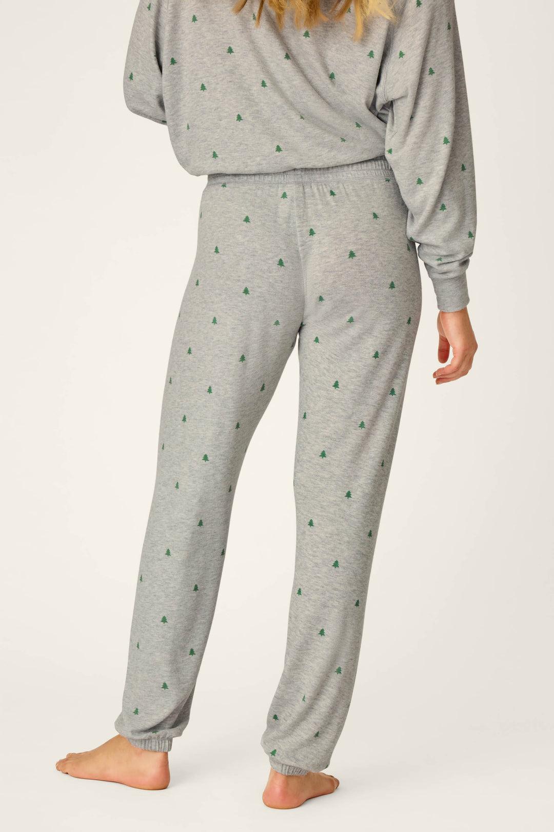 A person stands barefoot and facing away, wearing P.J. Salvage Happy Campawr Tree Print Jogger Pants in grey with small green tree patterns. The matching top is partially visible against a plain white background.