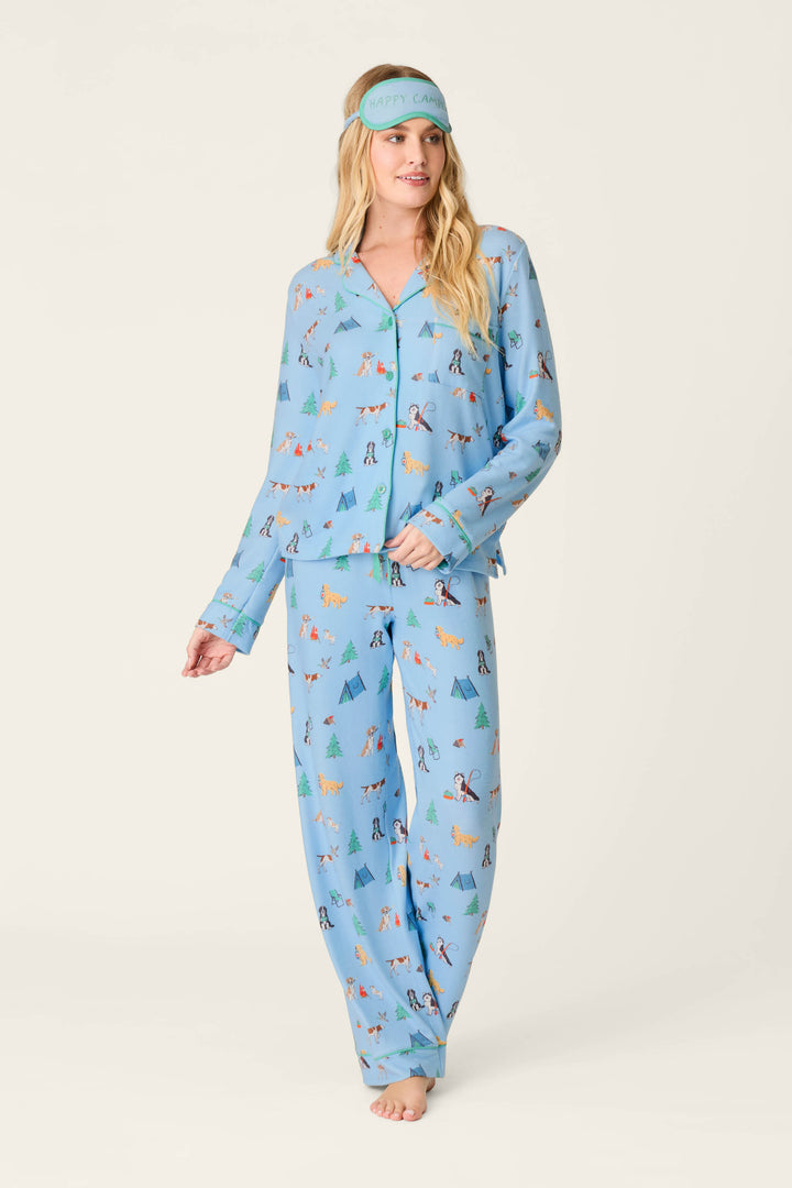 A woman wears the Happy Campawr Dogs Camping Print 3-Piece PJ Set by P.J. Salvage, featuring blue pajamas with playful camping dog and tree motifs, along with a matching blue sleep mask that reads “Happy Camper,” standing barefoot against a plain light background.