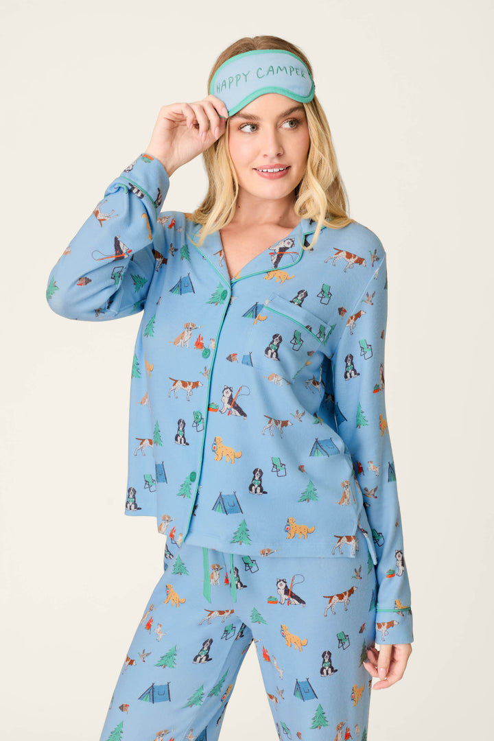 A woman dressed in the P.J. Salvage Happy Campawr Dogs Camping Print 3-Piece PJ Set, featuring blue pajamas with playful camping dog prints and a matching sleep mask reading “Happy Camper,” stands against a plain background, smiling as she lifts the mask just above her eyes.